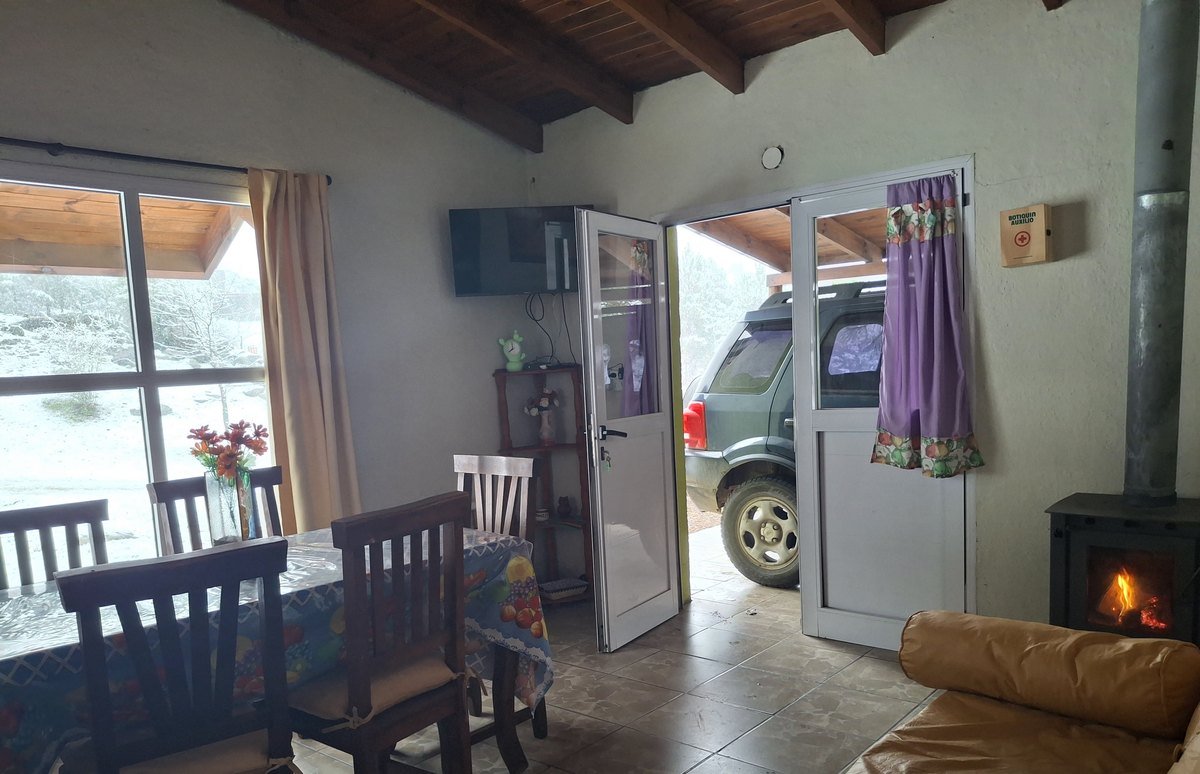 Living de la Cabaña Verde con la estufa a leña encendida y la nieve afuera, en Villa Yacanto